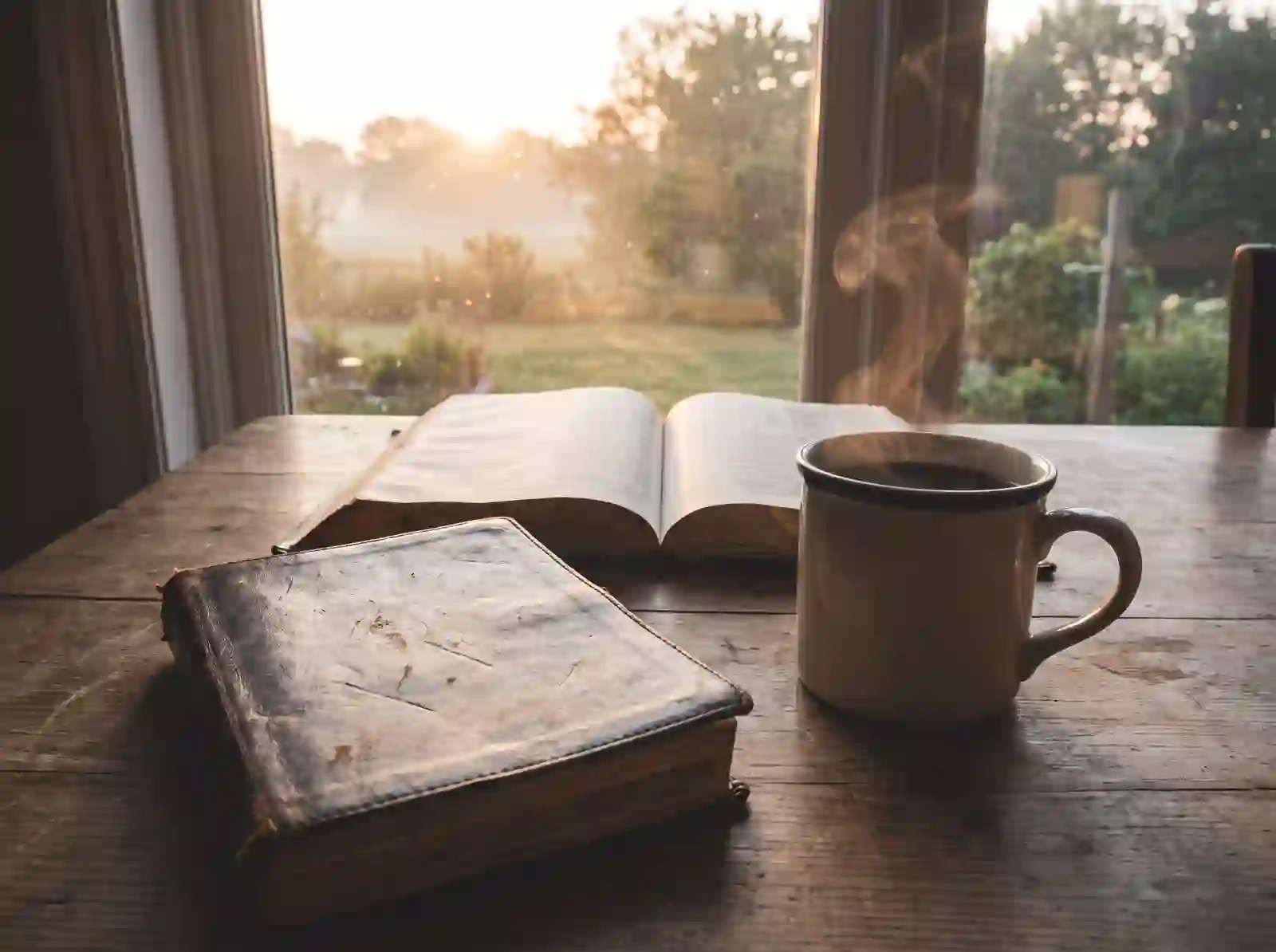 An open Bible, worn journal, and cup of coffee on a table at dawn